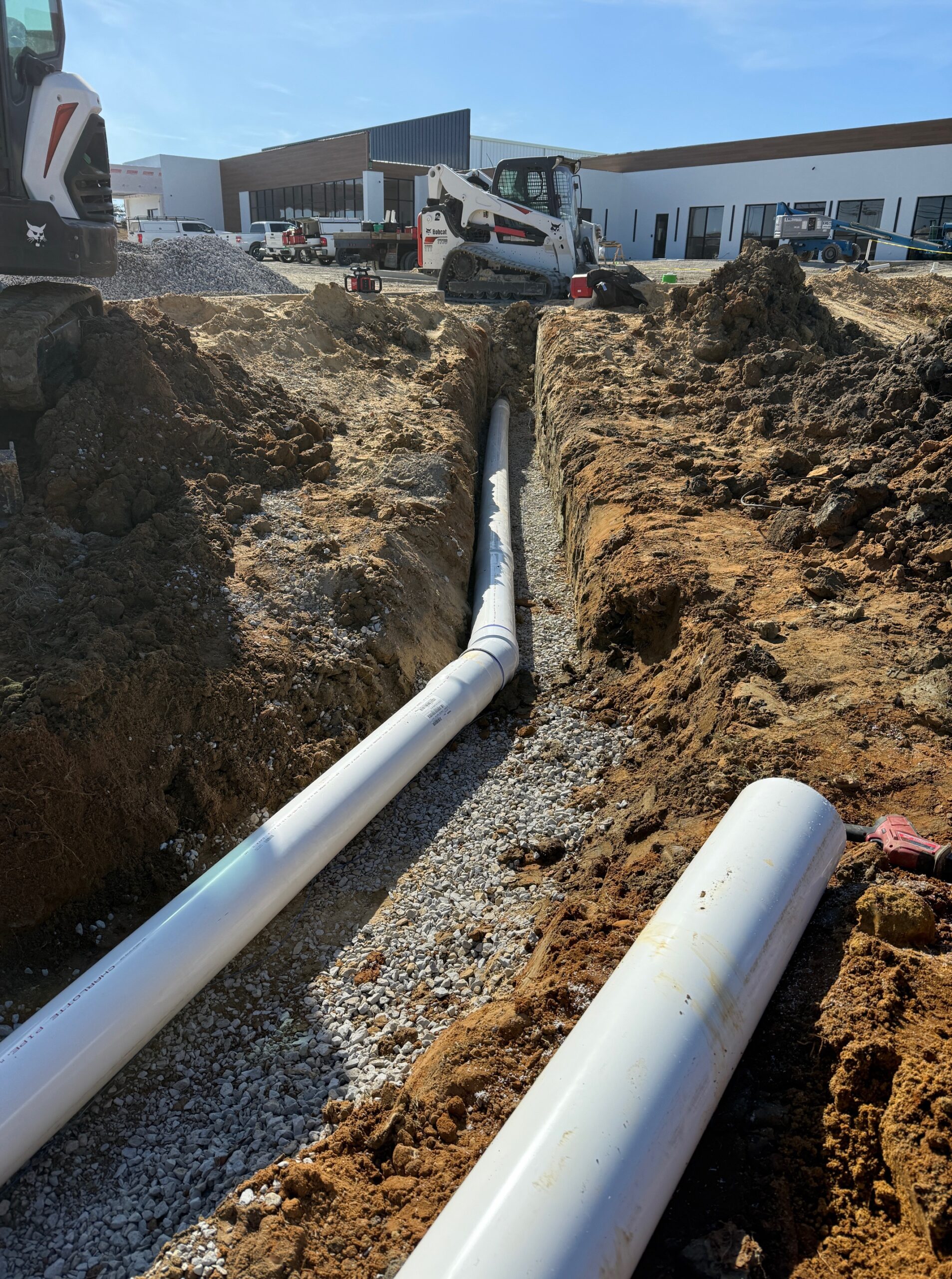 Large white PVC pipes laid in a gravel-bedded trench at a commercial construction site, with a Bobcat skid steer and modern building in the background.