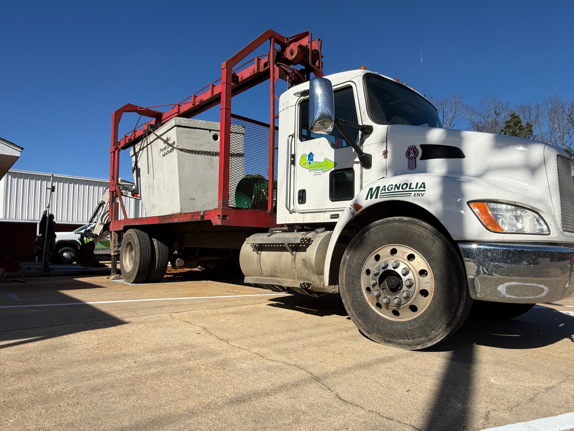 A white heavy-duty flatbed truck equipped with a red hydraulic crane lifting a large concrete septic tank in a parking lot on a clear, sunny day.