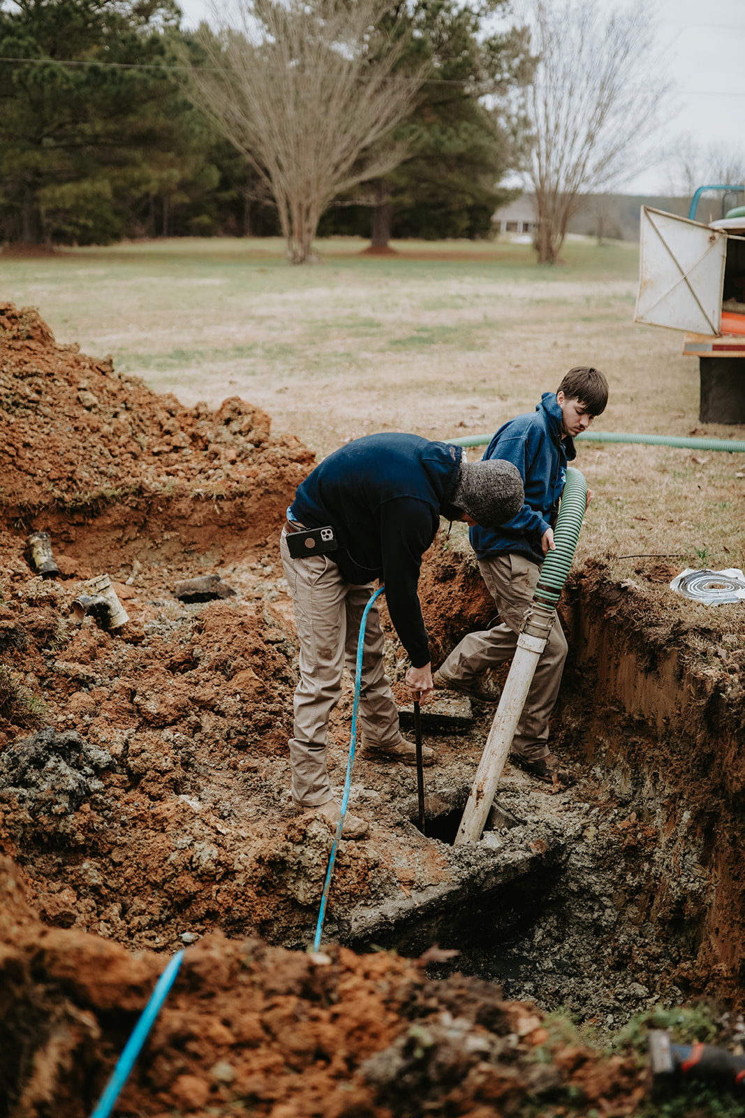 Two crew members working in an excavated dirt trench to install or repair white PVC septic lines and a green suction hose on a residential property.