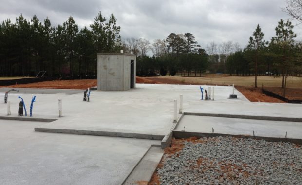A newly poured concrete building foundation featuring a central reinforced concrete storm shelter and various plumbing stubs, situated in a cleared area near a pine forest.